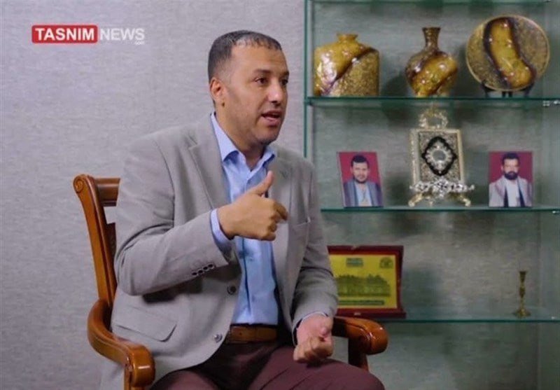A man in a gray blazer gestures while seated in a wooden chair during a Tasnim News interview, with decorative vases, framed photos, and a Quran displayed on shelves behind him.