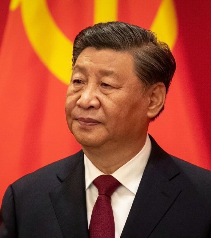 A man in a dark suit and red tie stands before a red flag with yellow stars, smiling slightly.