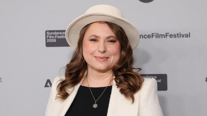 A woman wearing a white hat and blazer smiles in front of a step-and-repeat backdrop displaying "Sundance Film Festival 2026" logos.