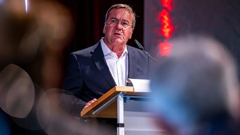A bespectacled man in a dark suit and white shirt speaks at a podium with a microphone, with blurred figures in the foreground.