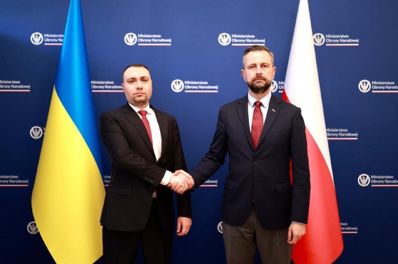 Two men in dark suits shake hands in front of the Ukrainian and Polish flags, with a backdrop displaying "Ministerstwo Obrony Narodowej" logos.