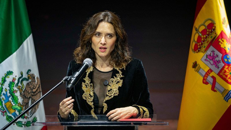 A woman speaks at a clear podium, flanked by the Mexican flag and the Spanish flag, gesturing with her right hand while wearing a black jacket with gold embroidery.