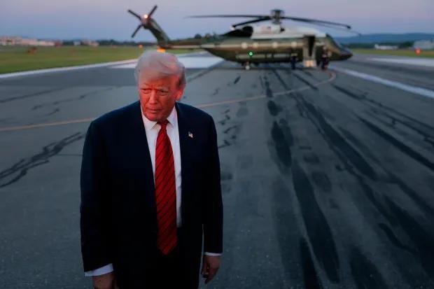A man in a dark suit and red tie walks across a tarmac, with a large helicopter visible in the background at dusk.