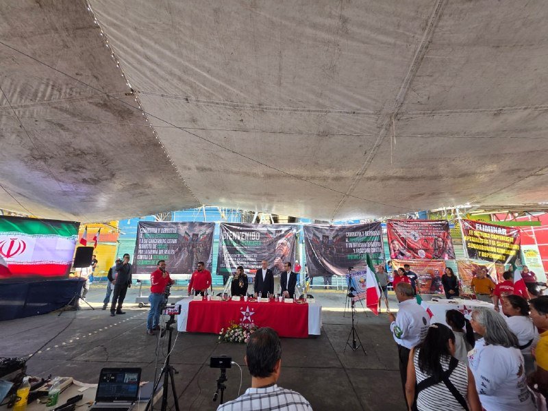 People gather under a large tent where several individuals stand behind a red-draped table, with banners reading "BIENVENIDO" displayed behind them and a Mexican flag visible.