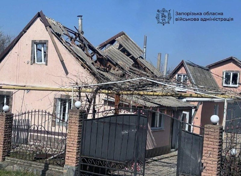 A pink residential house with a severely damaged, partially collapsed roof stands behind a closed metal gate, featuring an emblem and text identifying a regional military administration in the corner.