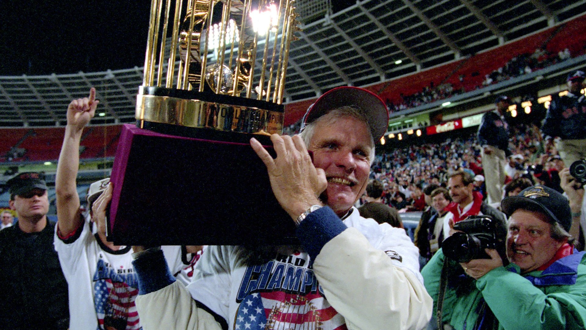 A smiling older man wearing a championship shirt hoists a large gold trophy on a stadium field, surrounded by cheering crowds and photographers.