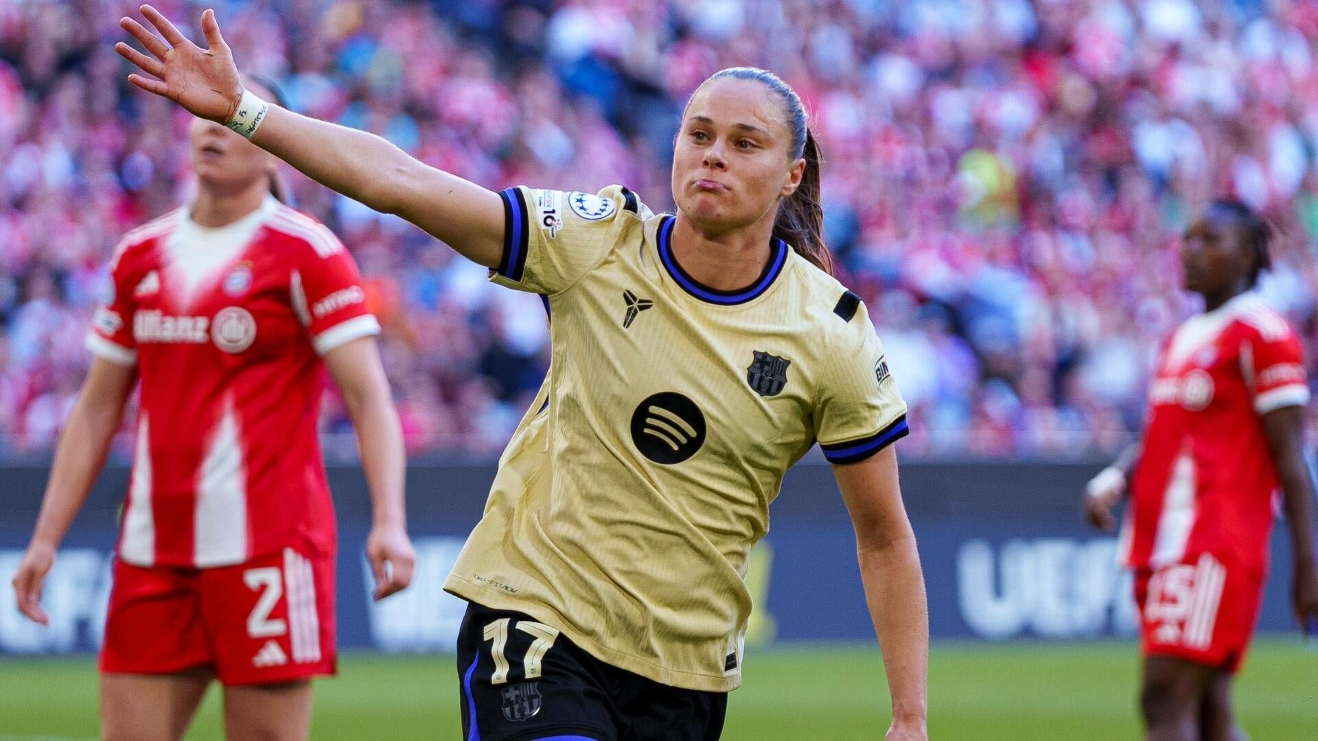 A female soccer player in a gold jersey raises her arm while standing on the field, with opposing players in red uniforms and a crowd visible in the background.