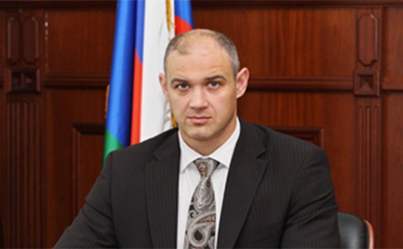 A man in a dark suit and patterned tie sits before a wooden paneled wall with a tricolor flag behind him.