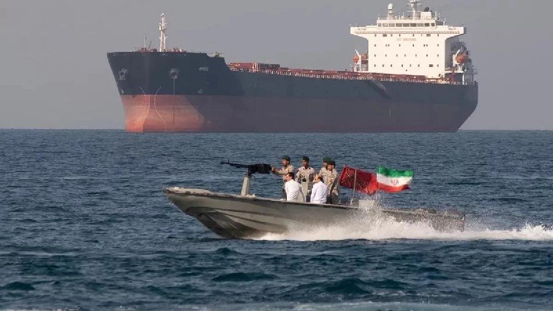 A small speedboat carrying armed personnel and flying an Iranian flag speeds across the water in front of a large cargo tanker on the open sea.