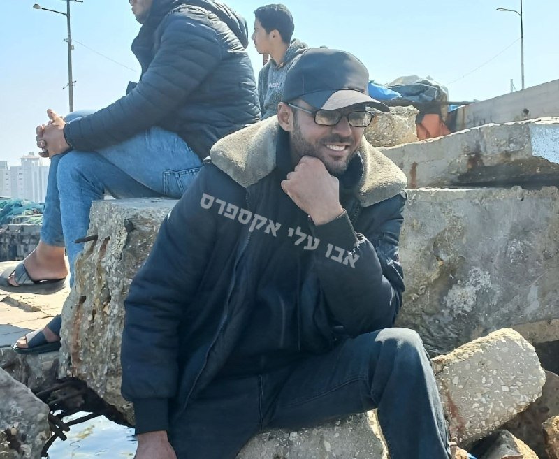 A smiling man wearing glasses, a black cap, and a dark jacket with Hebrew text sits on large concrete blocks near a waterfront, with other people seated behind him.