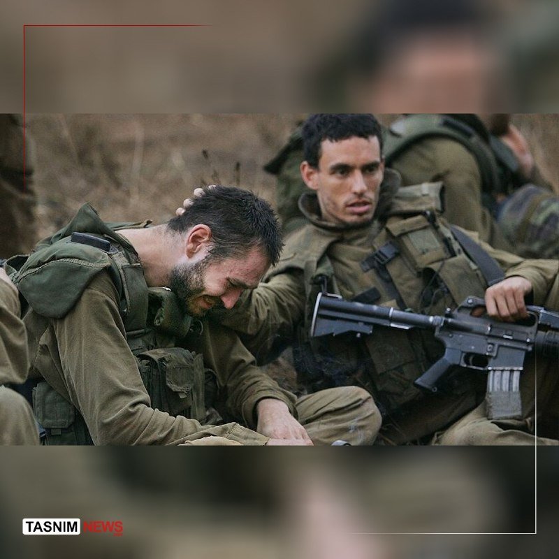 Two soldiers in olive-green uniforms and tactical gear sit outdoors; one bows his head while the other holds a rifle.