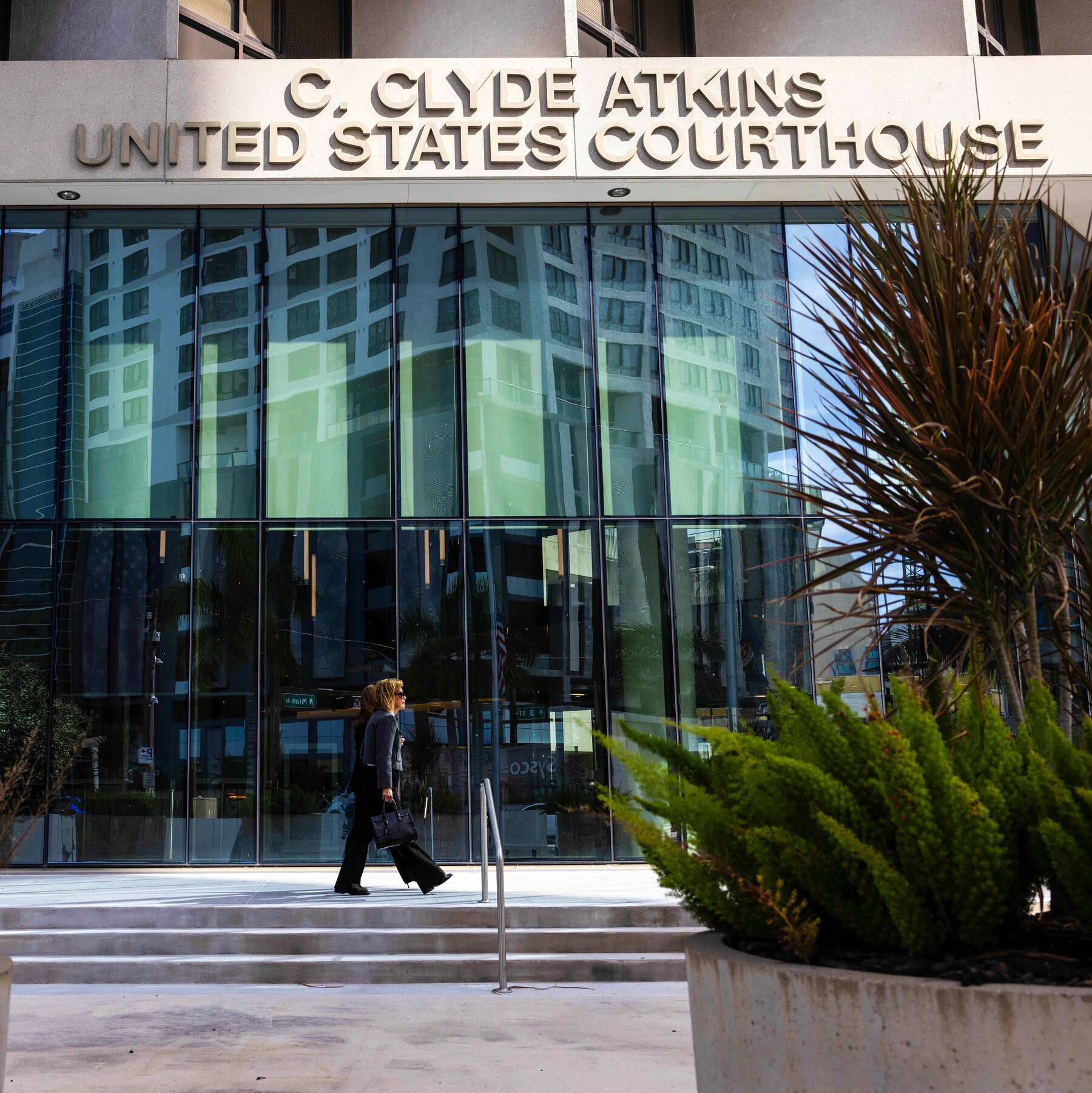 Two women carrying bags walk past the glass facade of the C. Clyde Atkins United States Courthouse.