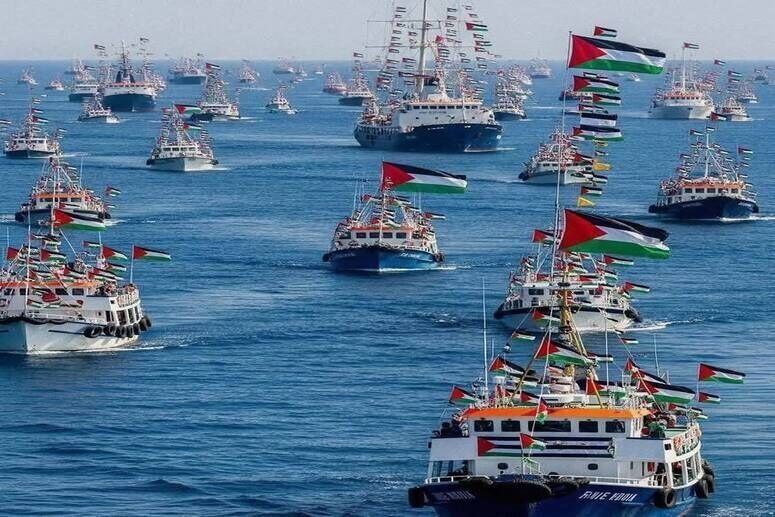 A large fleet of boats decorated with red, black, white, and green flags sails together across the ocean.
