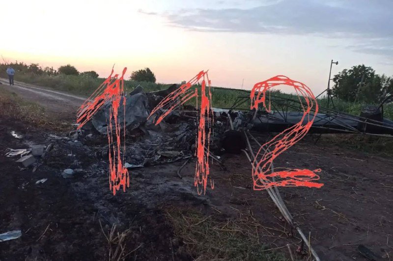Wrecked, burned debris sits along a rural road at dusk, with a figure standing in the distance.