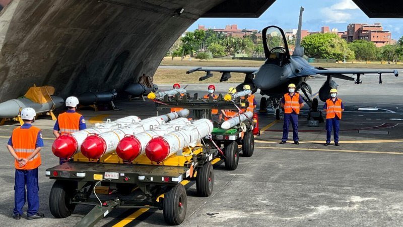 Ground crew in safety vests and helmets handle missiles on a tow cart near a fighter jet at an airfield.