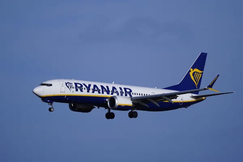 A blue and white Ryanair passenger jet with landing gear deployed is captured mid-flight against a clear blue sky, appearing to approach for landing.