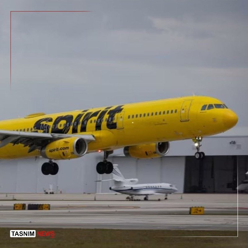A large yellow Spirit Airlines jet, landing gear extended, flies low over a runway as a smaller white private jet taxis below.
