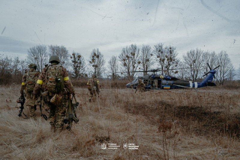 Armed soldiers in camouflage walk through a dry field toward a parked military helicopter under an overcast sky.