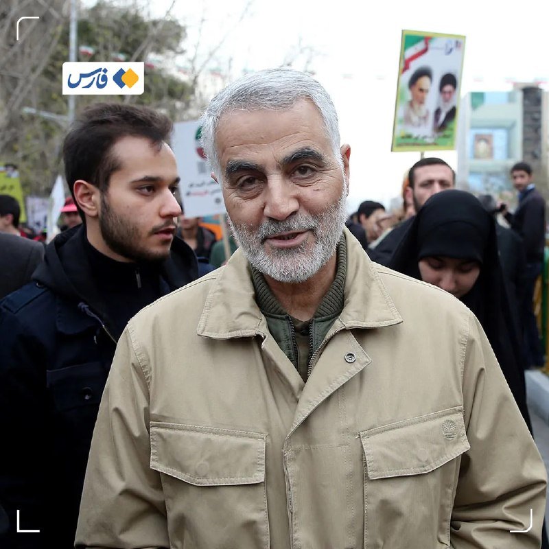 A gray-bearded older man in a tan jacket stands amid a crowd outdoors, with a poster bearing two portraits and an Iranian flag visible in the background.