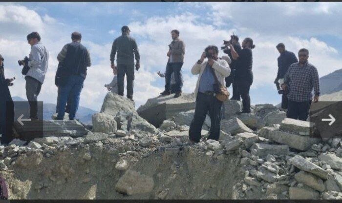A group of people, several holding cameras, stand atop a rocky, rubble-covered landscape under a partly cloudy sky with mountains in the distance.