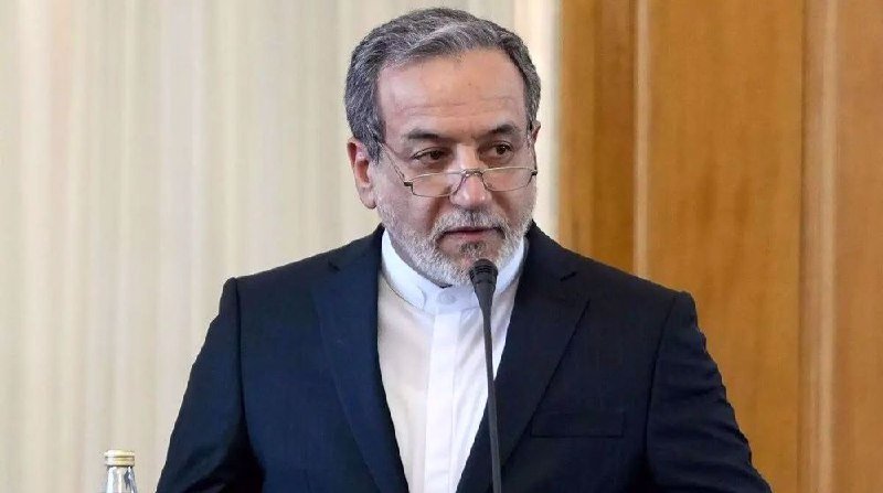 A gray-bearded man in a dark suit and white shirt speaks into a microphone during what appears to be a formal indoor event.