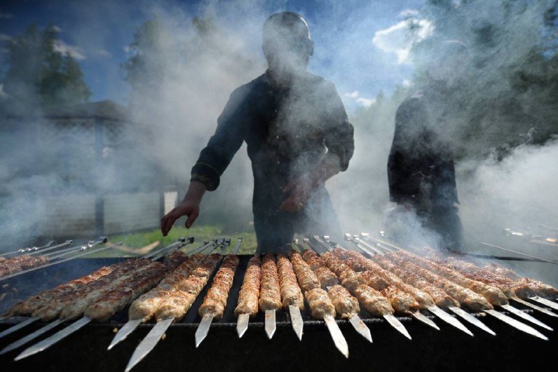 Two people stand behind a grill loaded with rows of skewered meat cooking amid heavy smoke under a blue sky.