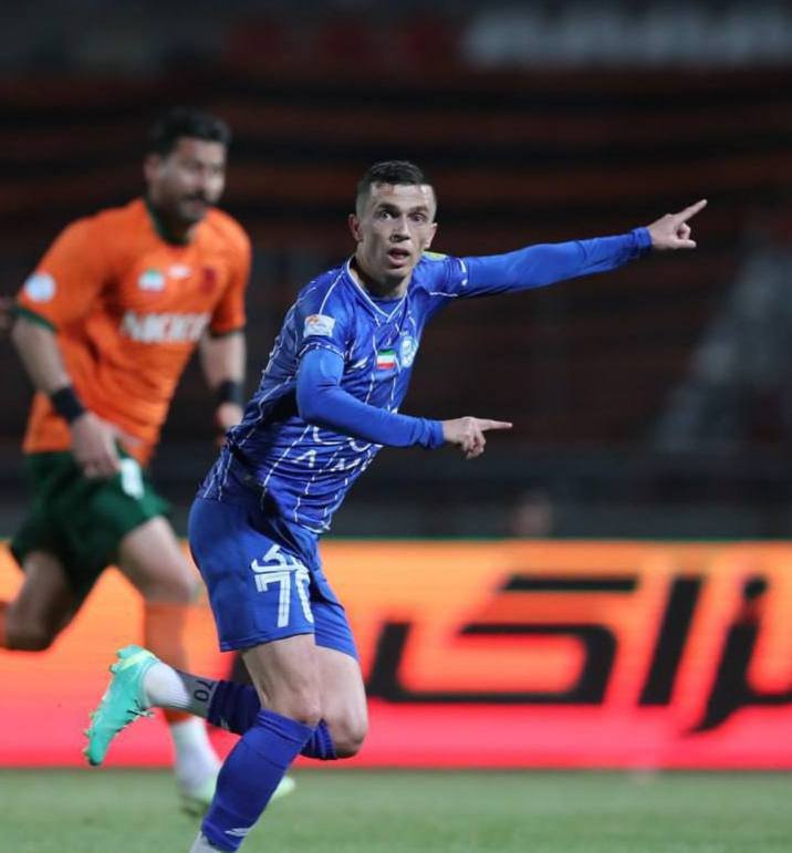 A soccer player in a blue uniform runs and points forward with his arm extended in celebration, while an opposing player in orange pursues him across the field.