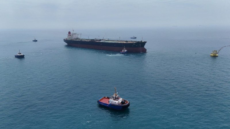 Aerial view of a large cargo tanker and several smaller tugboats navigating calm, open ocean waters under overcast skies.