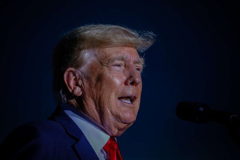A blond-haired man in a dark suit and red tie speaks into a microphone against a dark background.
