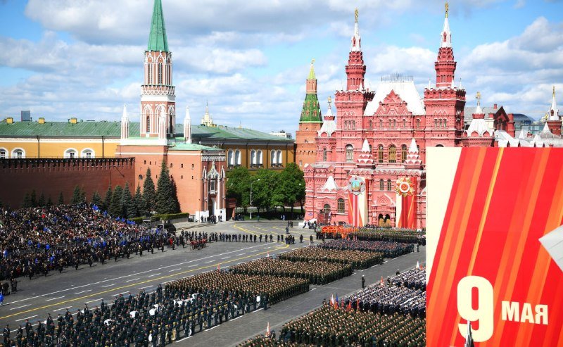 Military formations march in formation across Red Square during a parade, with the State Historical Museum and Kremlin walls visible behind them and a red "9 МАЯ" sign in the foreground.