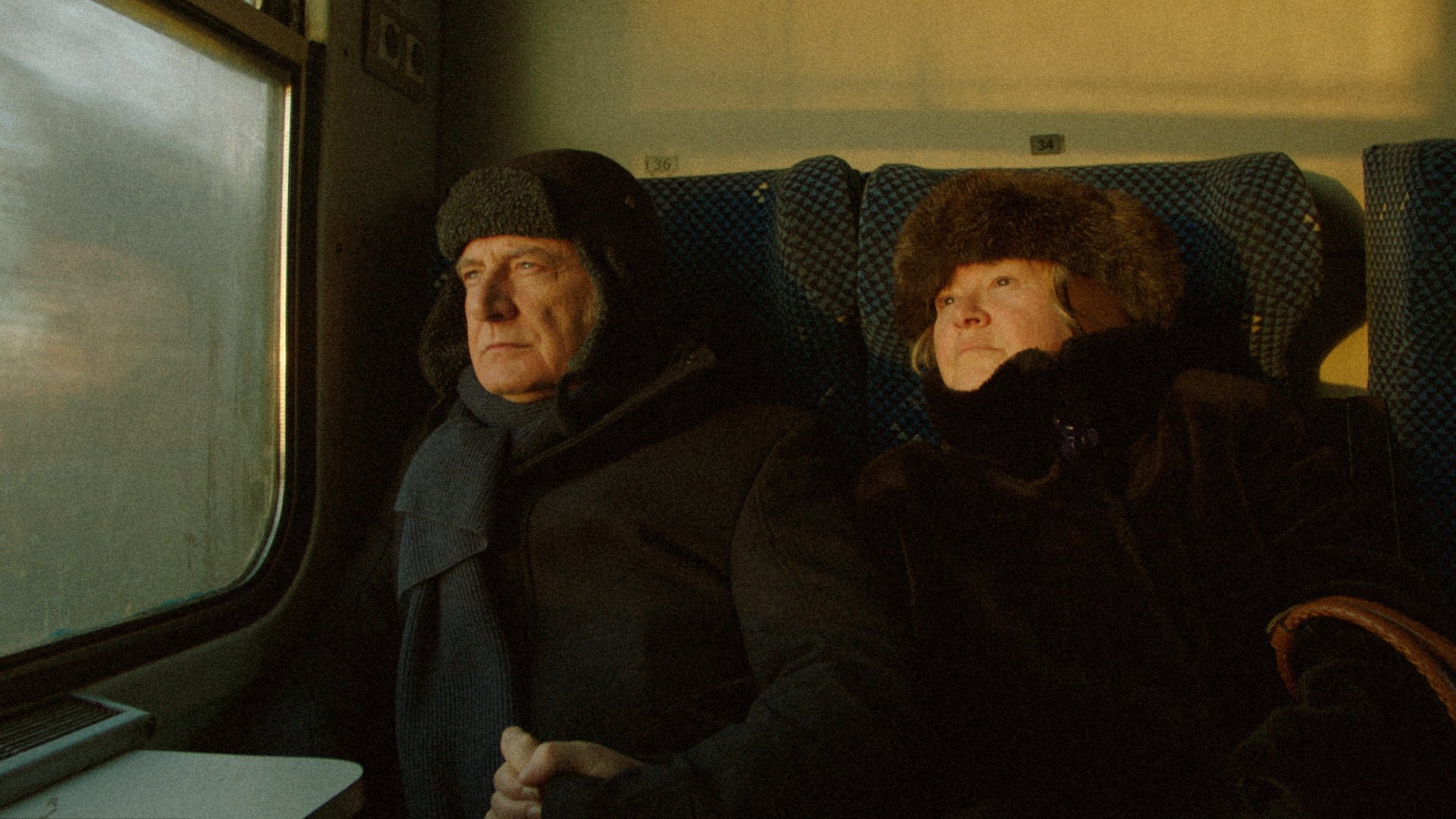 Two people in winter fur hats and dark coats sit beside a frosted train window on blue checkered seats.