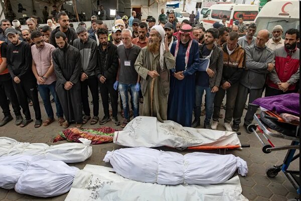 A group of men stand solemnly behind several white-shrouded bodies on stretchers and the ground in an outdoor urban setting, with ambulances visible in the background.