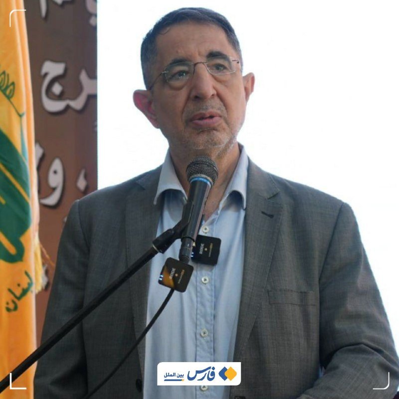 A bespectacled man in a gray suit speaks into a microphone, with a Persian-script banner and yellow flag visible behind him.