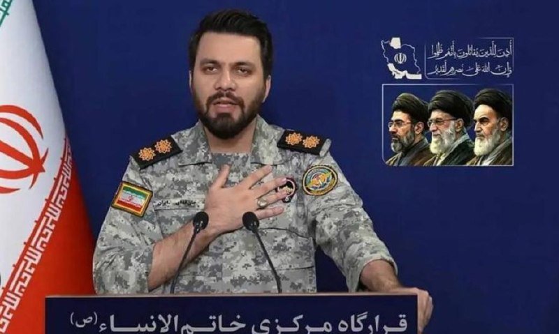 A bearded man in military camouflage uniform stands at a podium with Persian text, beside an Iranian flag, with three portraits displayed on the wall behind him.