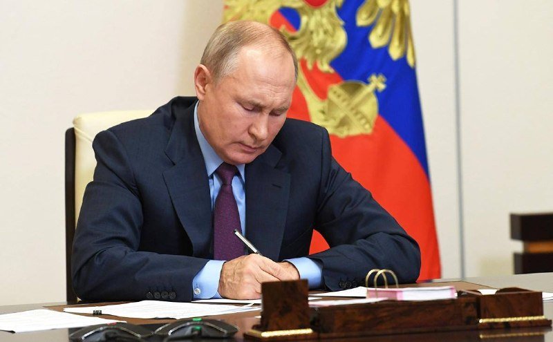 A man in a dark suit and burgundy tie sits at a desk, writing with a pen in front of a flag bearing a double-headed eagle emblem.
