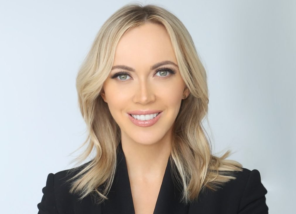 Blonde woman in a black blazer smiles at the camera against a plain light gray studio backdrop.