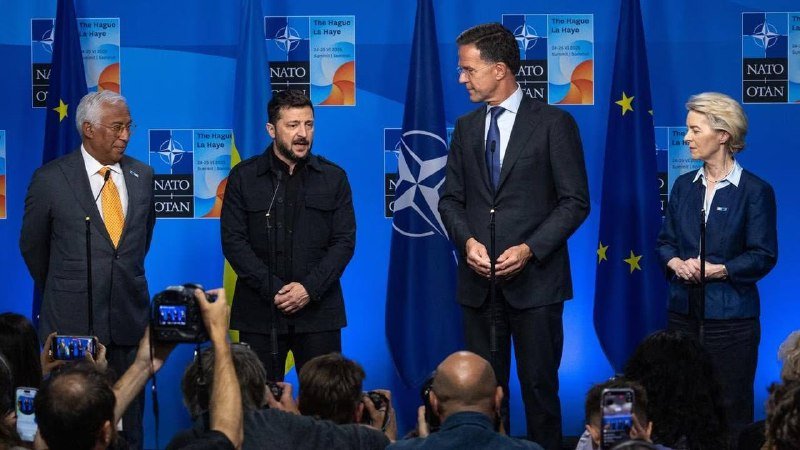Four officials stand at a podium before a blue backdrop featuring NATO and "The Hague La Haye" logos, while photographers take pictures from below.