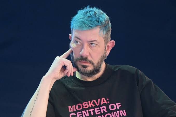 A man with gray hair and a beard, wearing a black t-shirt with pink text, touches his face while looking sideways against a dark background.