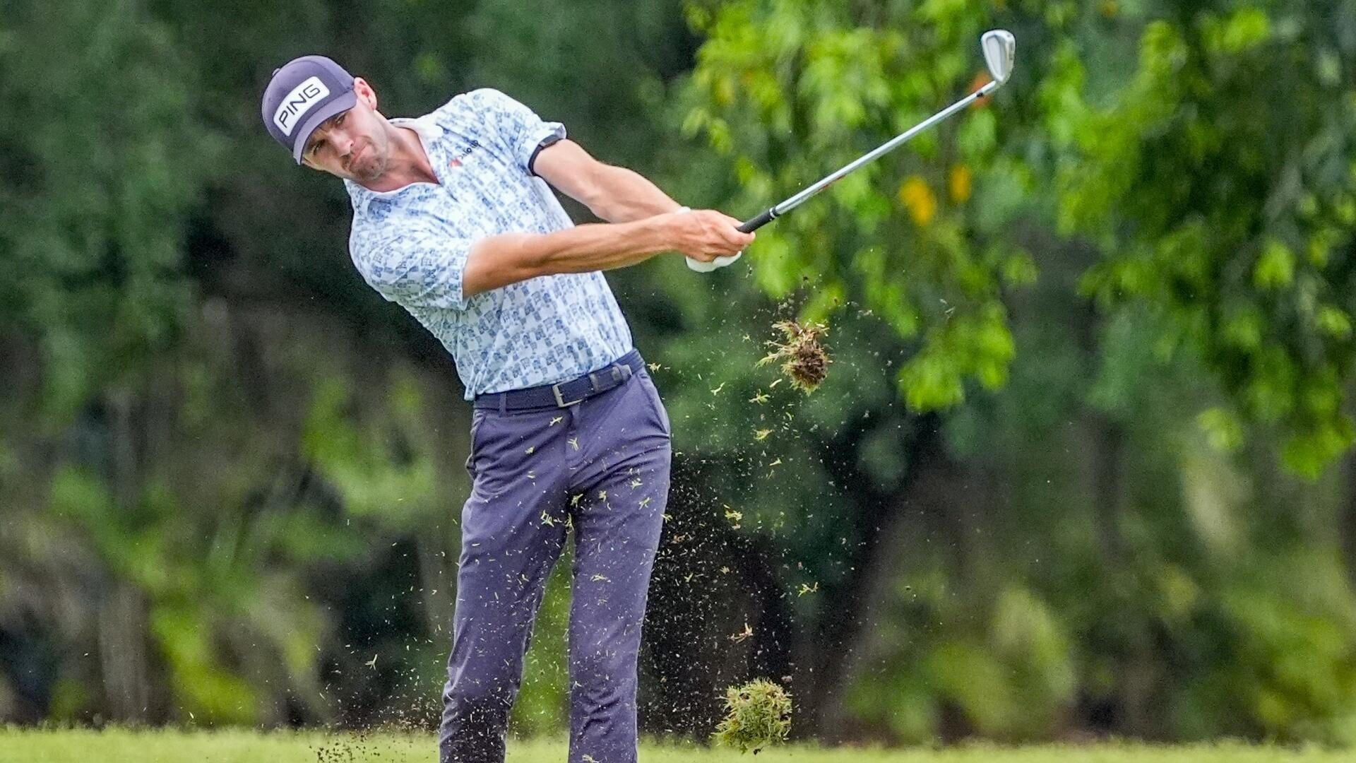 A golfer in a PING cap and patterned shirt swings an iron club from the rough, sending a clump of grass and dirt flying into the air against a backdrop of green trees.