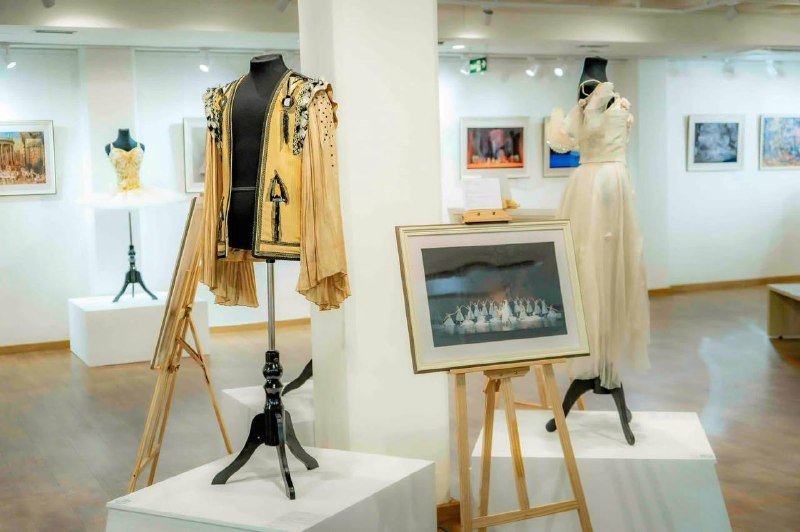 Two ornate costumes and a framed artwork rest on display easels in a gallery space with additional framed works visible on surrounding walls.