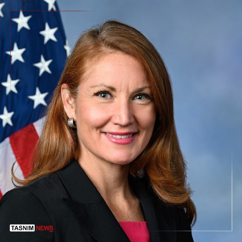 A woman with long auburn hair wearing a black blazer and pink top poses smiling in front of an American flag, with a "Tasnim News" watermark visible.