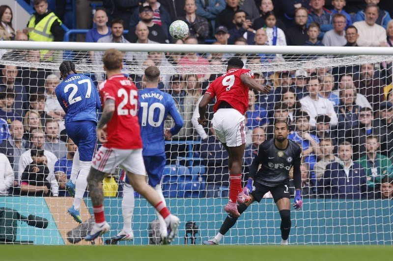 A player in a red jersey leaps to head a football near the goal during a match, as a goalkeeper in black and other players in blue jerseys look on.