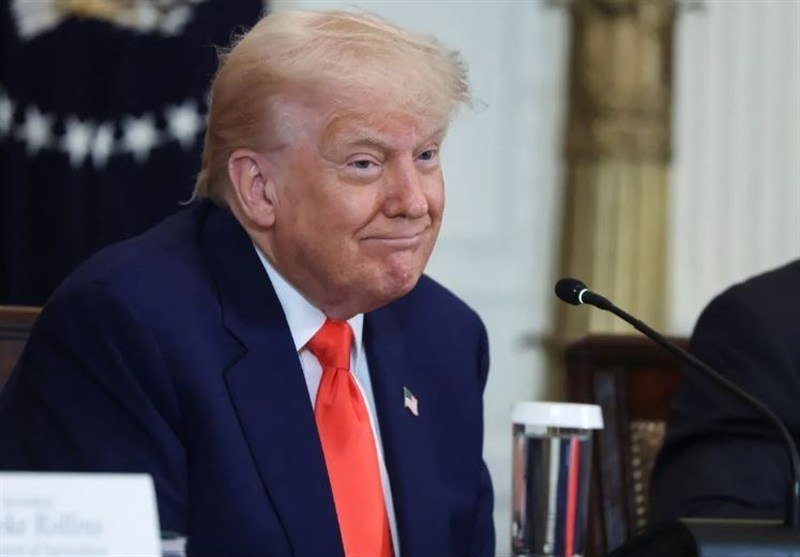 A blonde man in a dark suit and red tie smiles while seated at a table with a microphone and a striped tumbler.