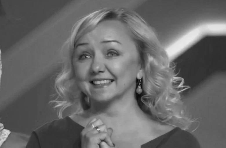 A smiling blonde woman wearing a dark top, pearl drop earrings, and rings looks upward in this black-and-white photo.