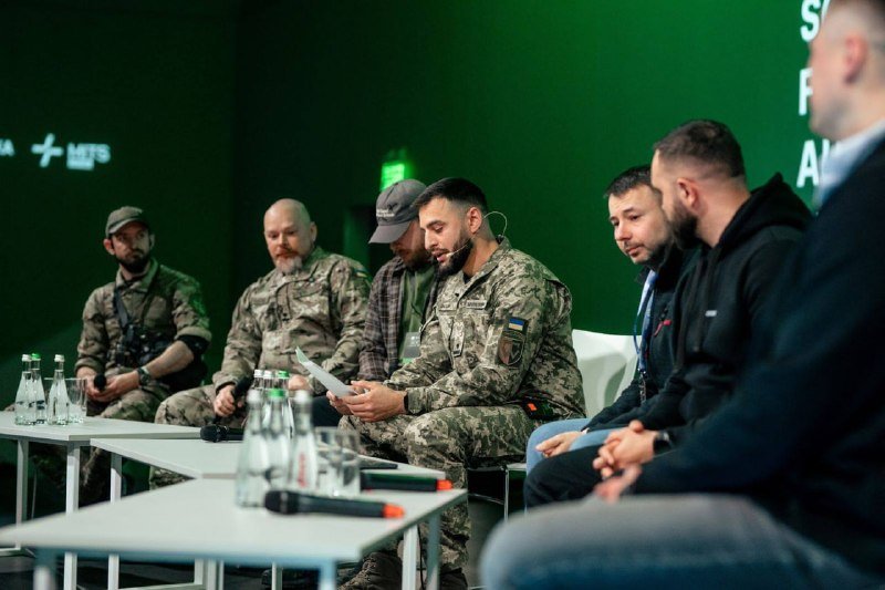 A panel of men in military camouflage uniforms and civilian clothing sit at a conference table reading papers against a green backdrop.