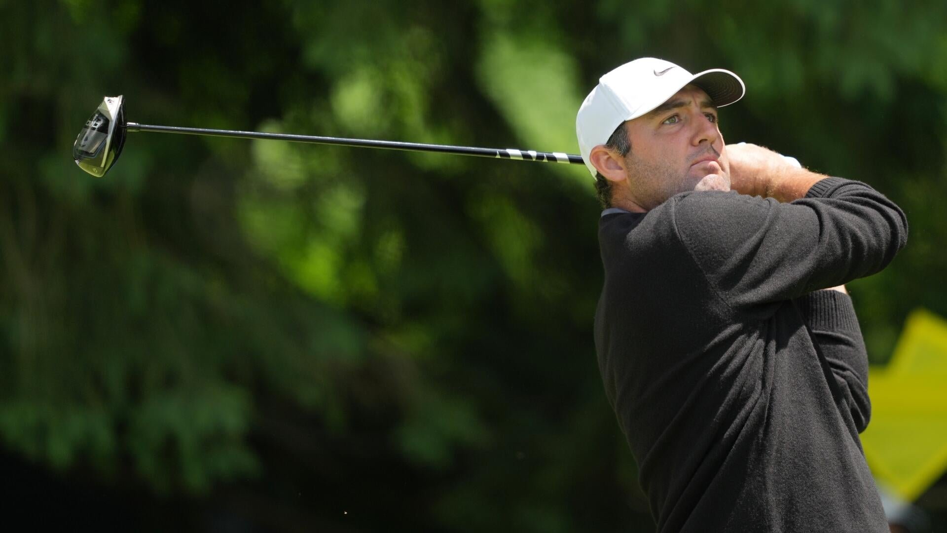 A golfer wearing a white cap and dark sweater completes a swing, eyes tracking the ball against a backdrop of green foliage.