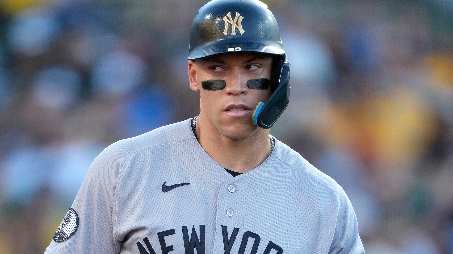 A baseball player wearing a gray New York Yankees jersey and a navy batting helmet with the number 99 stands in front of a blurred crowd.