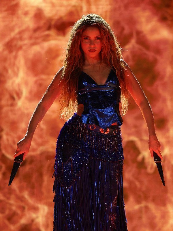 A woman with long curly red hair, wearing a blue sequined and fringed outfit, stands on stage holding two microphones against a fiery orange backdrop.