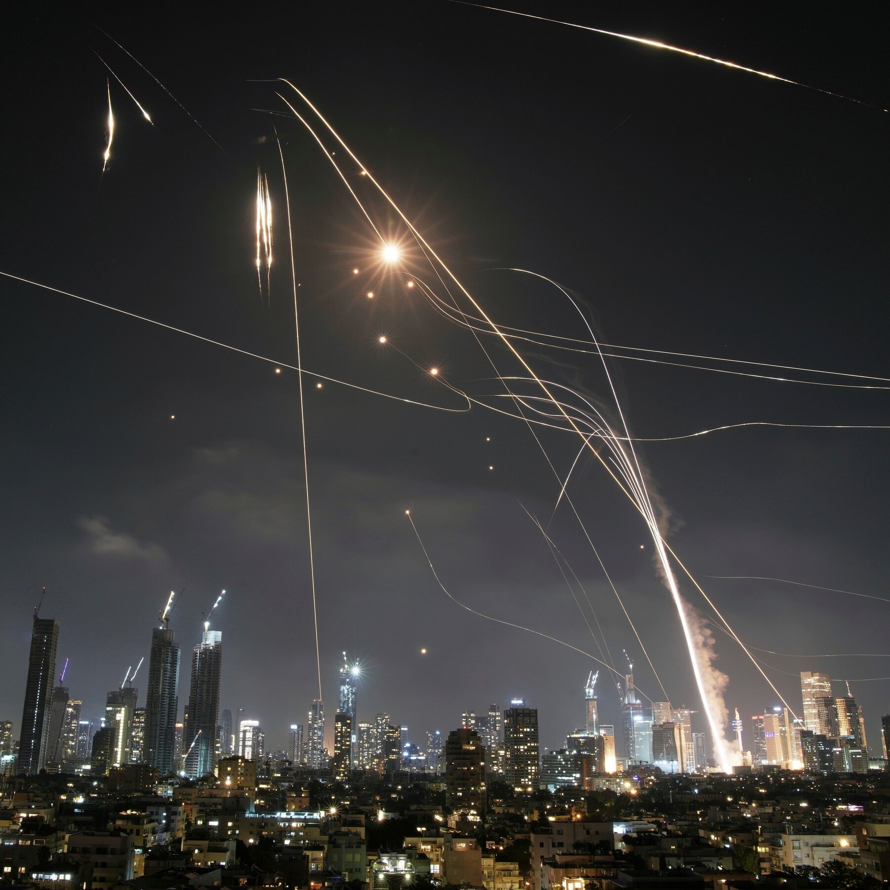 Bright projectile trails streak across the night sky above a lit urban skyline with high-rise buildings and construction cranes.