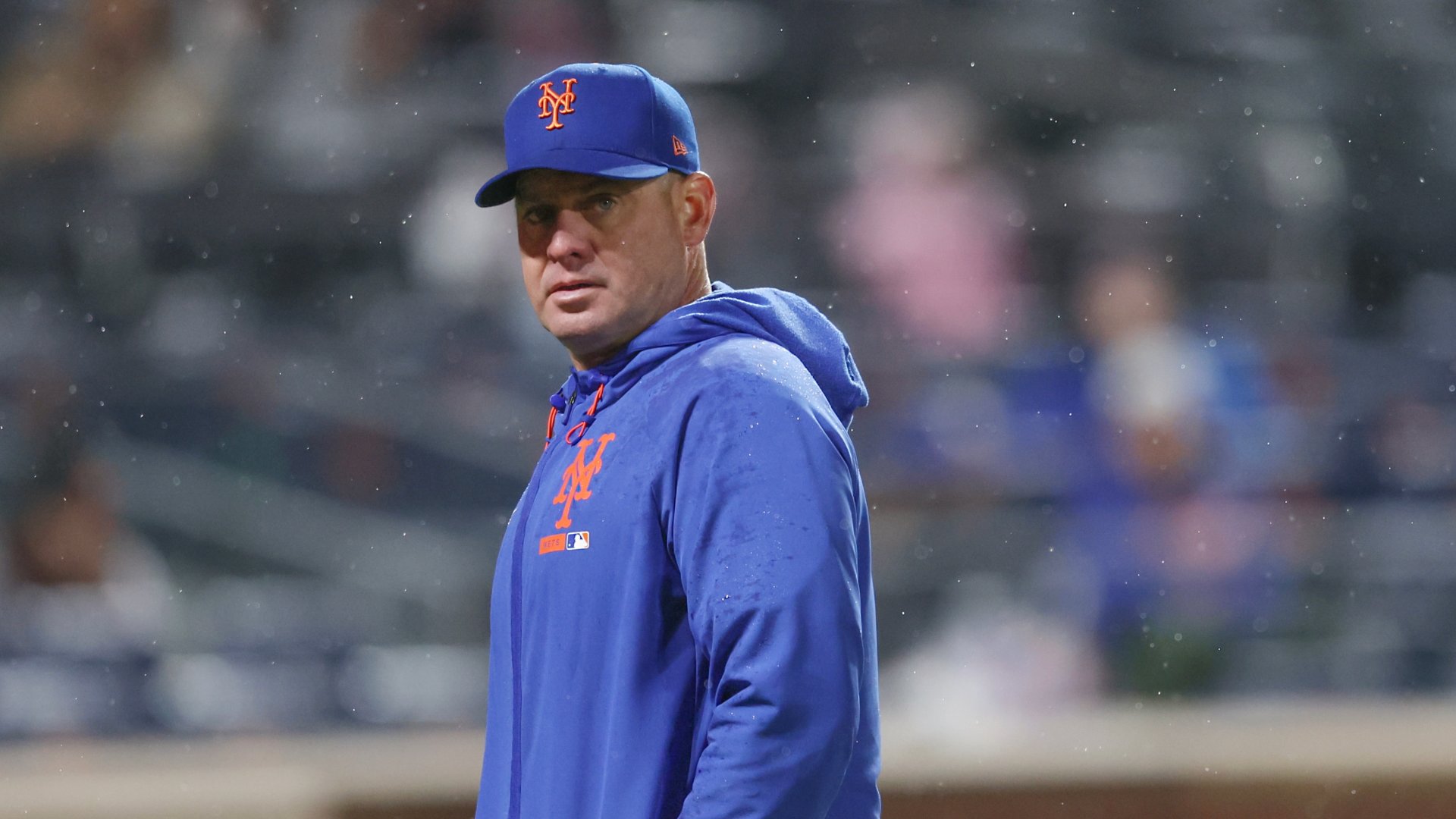 A man in a blue New York Mets cap and hoodie stands on a baseball field during a rainstorm, looking toward the dugout.